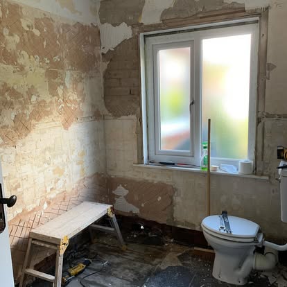 Bathroom mid-demolition with bare brick and adhesive-marked walls, new toilet installed, window, tools and step stool on dusty unfinished floor.