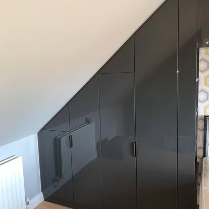Custom high-gloss dark grey wardrobes cut to fit under a sloped bedroom ceiling, with slim vertical handles beside a geometric headboard.