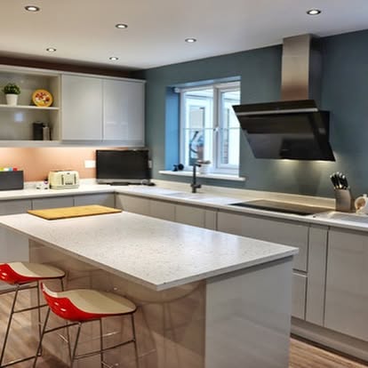 Sleek kitchen with light grey cabinetry, white speckled island, red-backed bar stools, teal accent wall, black range hood, and induction cooktop.