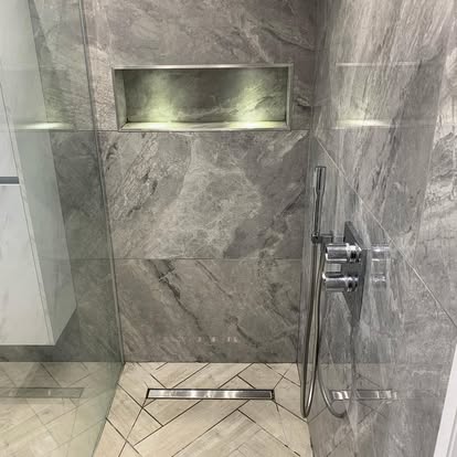 Walk-in shower with grey marble-effect wall tiles, LED-lit recessed niche, wood-effect chevron floor tiles, linear drain, and chrome fixtures.