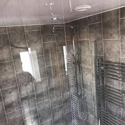Modern walk-in shower with large grey stone-effect wall tiles, dual chrome shower heads, frameless glass screen, and heated towel rail.