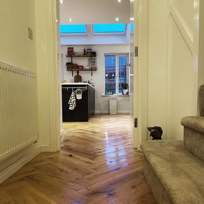 View from hallway showing light oak herringbone flooring leading into a modern kitchen with dark island, open shelves, skylights, and wall radiator.