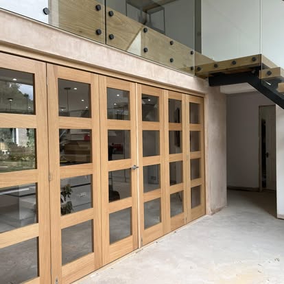 Light wood bi-fold doors with glass panels set in unpainted plaster walls above bare concrete floor, with mezzanine glass balustrade and staircase nearby.