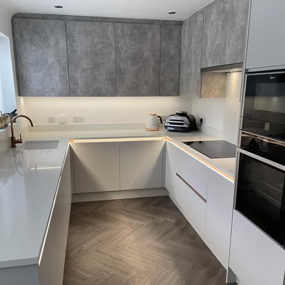 Modern U-shaped kitchen with grey textured uppers, white handleless lowers, white counters, copper faucet, herringbone wood floor, and black built-in appliances.