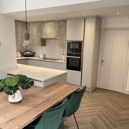 Bright open-plan kitchen and dining space with grey cabinetry, white counters, herringbone wood floor, induction hob, and green velvet dining chair.