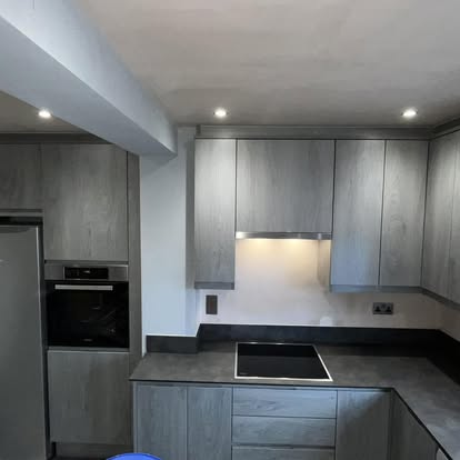 Minimalist kitchen with light grey vertical wood-grain cabinets, dark countertops, built-in black oven and cooktop, and under-cabinet LED lighting.