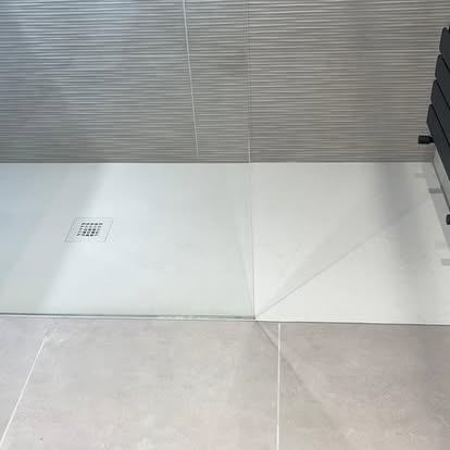 Close-up of low-profile white shower tray with linear drain, ribbed grey wall tiles, smooth grey floor tiles, and glass shower screen.