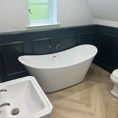 Attic bathroom with white freestanding oval bathtub against navy wainscoted wall, wall-mounted chrome taps, herringbone flooring, and small window.
