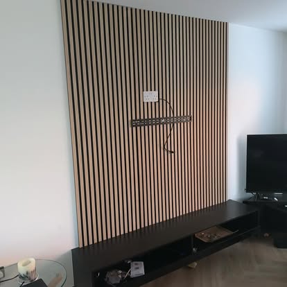Living room with vertical light wood slat accent panel above black floating media console, TV mount bracket, and herringbone wood flooring.
