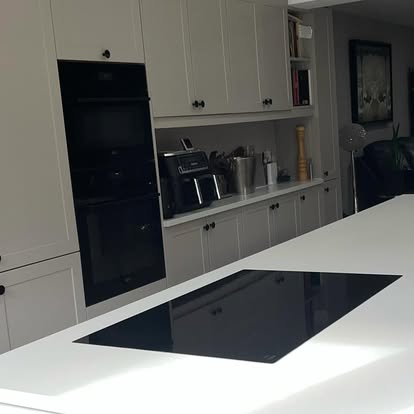 Contemporary kitchen with light grey shaker cabinets, black double wall oven, white island with integrated induction cooktop, and open shelving.