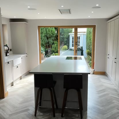Bright kitchen with white island and induction hob, grey shaker cabinets, farmhouse sink, herringbone floor, and timber bi-fold doors to the garden.