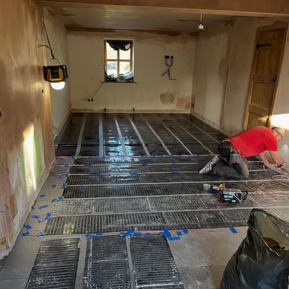 Renovation worker installing black electric underfloor heating mats on concrete subfloor in a plastered room with small window and wooden door.