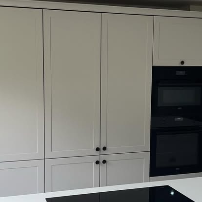 Light grey shaker kitchen cabinetry wall with black round knobs, integrated black double oven, and black glass cooktop on white counter.