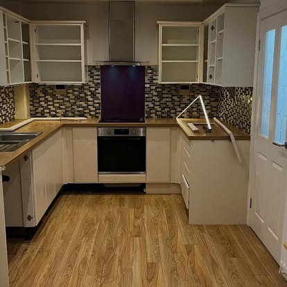 U-shaped kitchen mid-fit with cream cabinet carcasses, wood-effect countertop, mosaic tile backsplash, stainless oven and purple splashback panel.