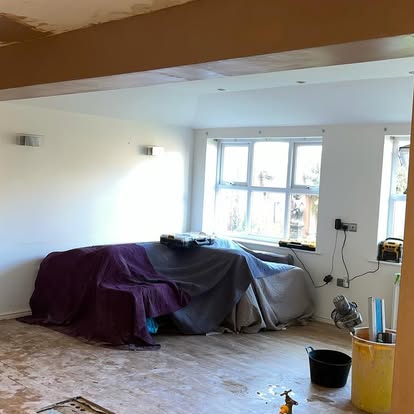 Bedroom work-in-progress with furniture covered in drop cloths, exposed wiring, unfinished plywood subfloor, and large bright window.