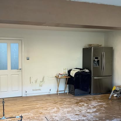 Kitchen area during remodel with stainless French-door refrigerator, dust-covered wood floor, glass-panel door, and exposed wall patches.