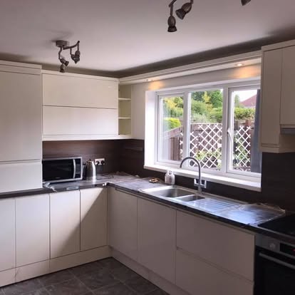 Modern L-shaped handleless cream kitchen with dark counters, wood backsplash, sink under a garden window, dark floor tiles, and protective wrap on the counter.