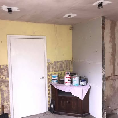 Room mid-renovation showing stripped yellow tiled wall, new grey drywall, white door, recessed spotlights with fresh plaster patches, and paint supplies.