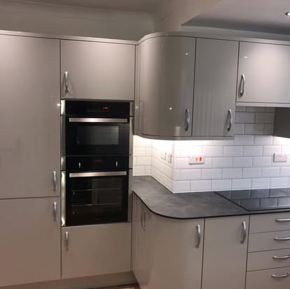 Modern kitchen corner with light grey high-gloss curved cabinets, dark matte countertop, white subway tile backsplash, under-cabinet lighting, and built-in double oven.