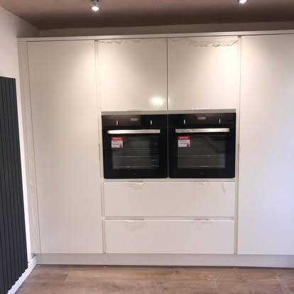 Floor-to-ceiling white kitchen wall with two integrated black ovens, tall pantry doors, designer radiator, and light wood-effect flooring.